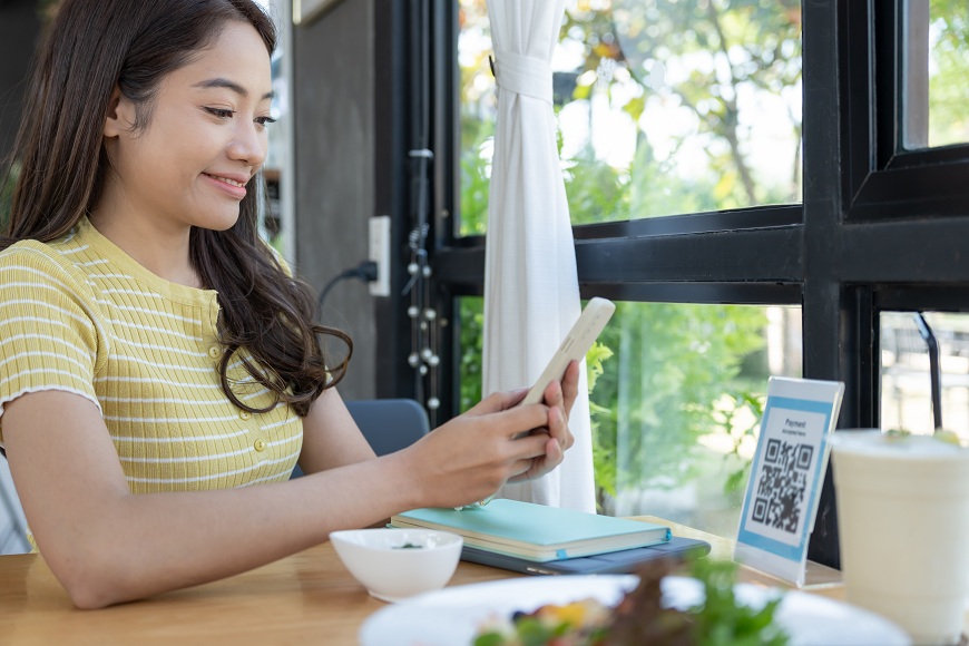 Jak platit mobilním telefonem? Žena sedí v restauraci a platí přes telefon pomocí QR kódu na stole.