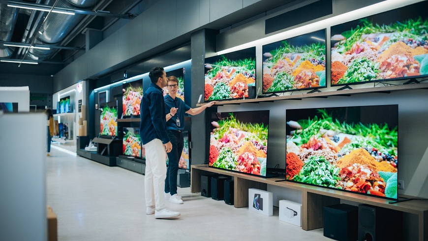 75, 98 nebo 115 palců? Rozdíl, který musíte vidět naživo! Muž stojí v prodejně před vystávkou televizí. Prodejce mu radí při výběru.